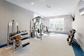 This single-floor fitness center is thoughtfully designed to provide a comprehensive workout experience across several distinct zones. The main strength training area features multiple cable and resistance machines, adjustable benches, and a full-length mirror wall that amplifies the sense of space while allowing users to monitor their form. Large windows along one wall invite abundant natural light, offering outdoor views that enhance the workout environment. A textured concrete-style accent wall adds an industrial character, contrasting with the clean grey and white color palette throughout. The flooring combines neutral carpet with bold blue accents that visually define different activity areas. A dedicated cardio and functional training zone showcases a rowing machine, medicine ball rack, and open floor space beneath a striking floor-to-ceiling forest mural, creating an immersive, nature-inspired atmosphere. Mounted flat-screen televisions are positioned throughout the facility for entertainment during workouts. A well-appointed refreshment and utility station features grey cabinetry, a beverage cooler, and counter space, providing added convenience. The facility is accessed through glass-paned doors and connects seamlessly between its zones, creating a cohesive and functional layout suited for diverse fitness routines.