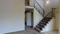 This two-story space is defined by its dramatic vertical proportions and open layout. The main living area immediately draws attention with its soaring ceiling height, creating an airy, expansive atmosphere. A tile-surround fireplace serves as a focal point on the main level, featuring classic white tile work and a traditional firebox. Neutral-toned carpet flows throughout the carpeted areas, complemented by tile flooring at the entryway transition points.

A prominent staircase with carpeted treads and sleek black metal railings rises from the main level to the upper floor, where an open-railing overlook provides a view down into the living space below, reinforcing the sense of height and connectivity between floors. Beneath the staircase, an alcove leads to additional rooms, making efficient use of the available space.

The walls throughout are finished in neutral tones, providing a clean, versatile backdrop. The overall design balances functional layout with architectural interest, highlighted by the double-height ceiling, open stair structure, and the warmth offered by the fireplace on the main level.