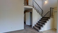 This two-story space is defined by its dramatic vertical proportions and open layout. The main living area immediately draws attention with its soaring ceiling height, creating an airy, expansive atmosphere. A tile-surround fireplace serves as a focal point on the main level, featuring classic white tile work and a traditional firebox. Neutral-toned carpet flows throughout the carpeted areas, complemented by tile flooring at the entryway transition points.

A prominent staircase with carpeted treads and sleek black metal railings rises from the main level to the upper floor, where an open-railing overlook provides a view down into the living space below, reinforcing the sense of height and connectivity between floors. Beneath the staircase, an alcove leads to additional rooms, making efficient use of the available space.

The walls throughout are finished in neutral tones, providing a clean, versatile backdrop. The overall design balances functional layout with architectural interest, highlighted by the double-height ceiling, open stair structure, and the warmth offered by the fireplace on the main level.