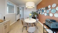 This charming café occupies a single, thoughtfully designed floor that balances functionality with warm, inviting aesthetics. The main seating area centers around a round white table surrounded by modern Eames-style chairs, positioned beneath a large, cylindrical drum pendant light that serves as the room's focal point. Natural light filters generously through wide windows fitted with white horizontal blinds, brightening the light tile flooring that runs throughout the space.

A standout accent wall in a rich terracotta tone adds warmth and visual interest, adorned with an artistic arrangement of decorative plates in varying shades of blue and white. Lush potted palms positioned throughout the café introduce a natural, organic element that complements the contemporary interior.

The beverage station along one wall features a granite countertop with a textured tile backsplash, equipped with a coffee maker and an assortment of refreshments, making it a practical and welcoming amenity for guests. Crown molding runs along the ceiling throughout, lending an elegant, finished quality to the interior.

Access is provided through a classic white paneled door with glass detailing, connecting seamlessly to adjacent spaces. A fire safety device is visibly installed, reflecting attention to building standards. The overall ambiance is clean, modern, and hospitable.