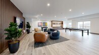 Located at 50 Otterbein Rd in Kitchener, Ontario, this single-floor amenity space offers a generously sized, open-concept layout designed for both relaxation and recreation. The interior is finished with wide-plank light hardwood flooring throughout, crisp white ceilings with recessed lighting, and clean neutral walls that create a bright, contemporary atmosphere.

The living area serves as a central focal point, anchored by a curved navy sectional sofa, a cognac leather lounge chair, and a round accent chair, all arranged on a textured area rug. A wall-mounted flat-screen TV is framed by a slatted wood accent wall, adding warmth and visual depth. Lush potted plants and bold, colorful artwork displayed on a built-in shelving unit contribute to the space's vibrant character.

The recreation zone is well-appointed, featuring a full-size pool table, a foosball table, and a shuffleboard table, catering to a variety of social activities. A built-in wooden bar or service counter provides additional functionality. Natural light flows in through large windows and a sliding glass door that opens to an outdoor terrace, offering outdoor views and enhancing the open, airy feel of the space. This thoughtfully designed lounge combines comfort, style, and entertainment in one cohesive environment.