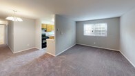 This single-floor residential unit offers a practical and comfortable layout designed for everyday living. The kitchen is equipped with warm honey-toned wood cabinetry, laminate countertops, a stainless steel sink, and a black gas range with overhead ventilation. It opens toward the living area, creating a connected flow between cooking and entertaining spaces. The living room is generously proportioned, featuring soft wall-to-wall carpeting in a neutral gray tone and a window that brings in natural light with views of the surrounding greenery. Adjacent to the living area, the dining space is highlighted by an elegant chandelier, adding a touch of classic character to the room. Multiple bedrooms carry the same neutral carpeting and soft gray wall palette, each benefiting from natural light provided by windowed walls with blinds. One bedroom features a double-door closet with a rod and shelf system, offering ample storage. The bathroom is clean and functional, presenting a full tub and shower combination with chrome fixtures, tile flooring in a neutral stone pattern, and a vanity with wood cabinetry. Throughout the unit, white trim, consistent neutral tones, and plush carpeting create a cohesive, move-in-ready atmosphere.