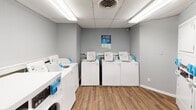 Located at 2222 Ashley River Rd in Charleston, SC, this single-floor laundry facility is designed for practical, efficient use. The space is finished with warm, wood-plank flooring and light gray walls throughout, creating a clean and consistent aesthetic.

The main laundry area is well-equipped with multiple rows of top-loading washing machines and front-loading dryers, all neatly arranged to maximize accessibility and workflow. A drop shelf along the wall provides a convenient folding surface. Overhead, a suspended drop ceiling with flush fluorescent lighting ensures bright, even illumination across the entire room. Signage promotes app-based payment and machine management, reflecting a modern, tech-enabled laundry experience.

The entry area features two exterior-facing doors — one with a glass panel and one designated as the emergency exit — alongside app-use instructional signage, offering a welcoming and informative transition into the facility.

An ADA-compliant restroom is accessible directly from the laundry area. It is equipped with grab bars, a standard toilet, a drop ceiling with ventilation, and matching hardwood-style flooring, ensuring comfort and accessibility for all users.

Overall, this facility presents a tidy, functional environment well-suited for shared or community laundry use in the Charleston area.