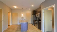 This single-floor space showcases a well-appointed open-concept kitchen that serves as the focal point of the layout. The kitchen is equipped with a full suite of stainless steel appliances, including a French door refrigerator, electric range, and built-in microwave. Dark espresso-toned cabinetry provides ample storage and creates a striking contrast against the granite countertop surfaces. A central kitchen island with matching granite offers additional workspace and features an integrated sink, with a stylish pendant light suspended above to provide focused task lighting. Recessed ceiling lights illuminate the space evenly throughout. Light-toned hardwood flooring runs throughout the area, adding warmth and cohesion to the overall aesthetic. The neutral wall tones complement both the cabinetry and flooring, creating a balanced and inviting atmosphere. Multiple doorways visible from the kitchen suggest convenient access to additional rooms, contributing to a functional and practical floor plan. The overall design balances modern finishes with a clean, understated aesthetic.