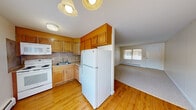 This single-floor, one-bedroom, one-bathroom unit offers a functional and well-proportioned layout designed to maximize every square foot. The combined kitchen and living area forms the heart of the unit, where the kitchen features warm honey-toned wood cabinetry extending to the upper walls, a tile backsplash, white appliances including a full-size refrigerator, electric range, built-in microwave, and a double sink — all set on light hardwood flooring. A large window in the kitchen floods the space with natural light. The living room is finished with neutral wall-to-wall carpeting, a chandelier, and a wide window with outdoor views, complemented by baseboard heating throughout. A separate dining nook with hardwood flooring and an elegant chandelier adds additional versatility to the common area. The bedroom is a comfortable, carpeted retreat featuring a built-in closet with louvered bifold doors and a window overlooking the exterior. The bathroom is tiled in classic cream, with a full bathtub and shower, a window for natural ventilation, and a vanity with a white toilet. Clean white walls and consistent baseboard heating are present throughout, giving the unit a cohesive and well-maintained character.