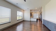 This single-floor unit showcases a clean, contemporary design with a thoughtfully planned open layout. The living area is spacious and bright, featuring rich hardwood-style flooring that extends throughout the main living spaces. Large windows fitted with horizontal blinds allow generous natural light to fill the room, offering views of the surrounding greenery and cityscape beyond.

The kitchen flows seamlessly from the living area and is defined by a peninsula counter that provides both workspace and a casual dining option. Gray shaker-style cabinetry is paired with white countertops and a textured stone tile backsplash, creating a modern yet warm aesthetic. Stainless steel appliances, including a refrigerator and dishwasher, complete the functional kitchen. Pendant lighting above the peninsula adds a refined touch.

A hallway connects the living spaces to the private areas of the unit. The bedroom features neutral carpet flooring and a bright window, offering a quiet and comfortable retreat. The bathroom is well-finished, presenting a dark-toned vanity with a white countertop, a large framed mirror, and a tiled tub-and-shower combination with an accent mosaic border. Light-toned tile flooring ties the bathroom together with an understated elegance throughout the unit.