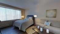This single-floor studio space features an open-concept layout that efficiently combines sleeping and living areas within one cohesive room. The design is anchored by dark hardwood flooring that runs throughout, providing a warm contrast against the light grey walls and white ceiling.

The sleeping area is centered around a queen-sized bed with a tufted navy headboard, dressed in neutral, soft bedding and complemented by a patterned area rug beneath. A pair of bedside lamps flank the bed, offering ambient lighting alongside the natural light that pours in through a large window dressed with vertical blinds. The window also features a built-in air conditioning unit beneath it.

The living area is defined by a cushioned grey loveseat accented with a decorative pillow, positioned alongside a dark wood coffee table. Decorative owl figurines and wall art add personality to the space. The layout maximizes the available footprint, making the room feel functional yet comfortable. The overall ambiance is calm and neutral, with a modern, understated aesthetic suited for everyday living.