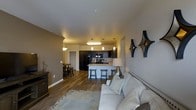 This single-floor space features an open-concept layout that seamlessly connects the living, dining, and kitchen areas, creating a cohesive and functional environment. The living area is anchored by a plush light-toned sofa, complemented by a patterned area rug over rich hardwood-style flooring that extends throughout the space. Neutral gray and warm beige wall tones provide a modern yet inviting backdrop, accented by decorative wall art.

The kitchen is a standout feature, showcasing dark espresso cabinetry paired with black appliances, including a refrigerator and range. A peninsula-style breakfast bar with bar stools serves as both a functional workspace and a casual dining spot. Pendant lighting above the bar adds a stylish touch, while additional overhead lighting ensures the kitchen is well-lit.

A separate dining area is visible toward the back of the space, furnished with a bistro-style table and chairs, ideal for more formal meals. The overall ambiance is contemporary and well-appointed, with consistent flooring and cohesive design elements tying each zone of the space together harmoniously.