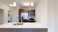 Located at 550 Halstead Avenue in Harrison, NY, this well-appointed one-bedroom, one-bathroom residence offers a thoughtfully designed single-floor layout with modern finishes throughout. Light hardwood flooring extends across the open living areas, creating a bright and cohesive feel.

The open-concept kitchen serves as a central feature, anchored by a large island with a quartz countertop, undermount sink, and dark wood cabinetry. Stainless steel appliances include a French door refrigerator with ice and water dispenser, an electric range, and an over-the-range microwave. Pendant and flush-mount lighting adds a polished touch. A stacked in-unit washer and dryer is conveniently tucked adjacent to the kitchen.

The living area is open and airy with clean white walls and ample room for flexible furniture arrangements. The bedroom features a large window that welcomes natural light and outdoor views, complemented by the same warm hardwood flooring.

The bathroom is finished to a high standard, featuring a dual-sink dark espresso vanity with white countertops, backlit mirrors, large-format tile walls, and a walk-in shower with a built-in grab bar—combining both style and accessibility. The overall ambiance is clean, contemporary, and move-in ready.