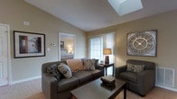 This single-floor residence offers a thoughtfully designed layout with a warm, inviting ambiance throughout. The living area serves as the heart of the home, highlighted by a striking skylight that floods the space with natural light, creating an airy and open atmosphere. Neutral beige walls and soft carpeted flooring contribute to the room's comfortable, understated elegance.

The living room is well-appointed, featuring a generous sofa accented with decorative throw pillows and a coordinating armchair arranged around a dark wood coffee table. A large window dressed with sheer curtains further enhances the natural light, while tasteful wall décor adds a refined finishing touch to the space.

The open doorway from the living area provides a seamless connection to the bedroom, which is visible in the background and continues the same warm, neutral color palette. The bedroom appears comfortably sized, illuminated by soft ambient lighting.

Overall, this residence combines practical layout with architectural details — most notably the skylight — to create a bright, welcoming living environment. The cohesive design language across rooms, characterized by neutral tones, soft textures, and carefully considered furnishings, gives the space a harmonious and livable character throughout.