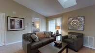 This single-floor residence offers a thoughtfully designed layout with a warm, inviting ambiance throughout. The living area serves as the heart of the home, highlighted by a striking skylight that floods the space with natural light, creating an airy and open atmosphere. Neutral beige walls and soft carpeted flooring contribute to the room's comfortable, understated elegance.

The living room is well-appointed, featuring a generous sofa accented with decorative throw pillows and a coordinating armchair arranged around a dark wood coffee table. A large window dressed with sheer curtains further enhances the natural light, while tasteful wall décor adds a refined finishing touch to the space.

The open doorway from the living area provides a seamless connection to the bedroom, which is visible in the background and continues the same warm, neutral color palette. The bedroom appears comfortably sized, illuminated by soft ambient lighting.

Overall, this residence combines practical layout with architectural details — most notably the skylight — to create a bright, welcoming living environment. The cohesive design language across rooms, characterized by neutral tones, soft textures, and carefully considered furnishings, gives the space a harmonious and livable character throughout.