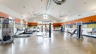 Located at 1562 Magnolia Dr in Oxford, Ohio, this expansive fitness center offers a generously sized, single-floor layout designed to accommodate a full range of workout activities. The space is immediately defined by its striking vaulted ceilings, which rise dramatically overhead and are serviced by exposed industrial-style ductwork and large paddle ceiling fans, ensuring optimal airflow throughout. Bold horizontal bands of orange, navy, olive, and black line the walls, lending the facility a vibrant, energetic atmosphere.

Natural light pours in through numerous windows and glass entry doors positioned along multiple walls, creating a bright and welcoming environment. The polished concrete flooring throughout the main area transitions to rubber matting in designated strength zones, providing both durability and safety. A dedicated free weights area features a full dumbbell rack, adjustable Matrix benches, and a power rack with plate storage, while a separate zone houses cable machines, a functional trainer, and a rowing machine. Cardio equipment, including multiple treadmills and upright machines, lines one side of the floor.

Additional amenities include TRX suspension straps, a heavy punching bag, stability balls, a wall-mounted flat-screen television, wall mirrors, and a drinking fountain. Multiple entry points and ample open floor space allow for comfortable movement and group exercise, making this fitness center a thorough and well-appointed facility.