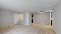 This single-floor space presents an open, functional layout finished in soft neutral tones throughout. The main living area is finished with light beige wall-to-wall carpeting, which adds warmth underfoot and complements the cool gray painted walls. A ceiling fan with a brushed nickel fixture is mounted overhead, providing both air circulation and ambient lighting to the space.

The layout connects the living area to an adjacent section, separated by a partial wall that defines each zone without fully enclosing them. This semi-open configuration allows for a natural flow between the living and dining or secondary living areas. A doorway along the partial wall leads to the kitchen, which features dark cabinetry, light-toned countertops, and tile backsplash detail visible from the main area.

Near the entrance, a white front door is set alongside a pass-through window or niche in the wall, adding a subtle architectural detail. A second interior door provides access to an additional room. White trim and baseboards run consistently throughout, offering a clean contrast against the gray walls and creating a cohesive, tidy aesthetic across the space.