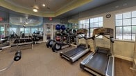 This fitness center is a thoughtfully designed single-floor facility offering a comprehensive workout environment for users of all fitness levels. The space is finished with neutral-toned carpet flooring and features elegant crown molding along the upper walls, which are painted in a calming two-tone palette of soft blue and yellow. Recessed lighting throughout ensures the space is well-lit, complemented by natural light streaming in through multiple windows fitted with blinds.

Floor-to-ceiling mirrors line the walls, visually expanding the space and allowing users to monitor their form during workouts. Multiple wall-mounted flat-screen televisions are strategically positioned throughout the room for entertainment during exercise sessions.

The cardio section is well-appointed with several treadmills and stationary bikes, while the strength training area features a diverse array of cable machines, weight benches, and selectorized strength equipment suitable for full-body training. A dedicated free weights section includes dumbbells and a medicine ball rack stocked with various weighted balls. Glass-panel double doors connect the fitness area to an adjacent space, adding an open, airy feel. The overall ambiance is clean, professional, and inviting, making this fitness center a functional and motivating space for daily exercise.