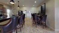 This single-floor space combines multiple functional areas into a cohesive, classically styled environment. The kitchen is equipped with stainless steel appliances, warm-toned wood cabinetry topped with decorative greenery, and dark granite countertops. A curved bar counter with studded leather swivel barstools provides casual seating, while industrial-style pendant lights suspended from the ceiling add a refined yet relaxed aesthetic. A wall-mounted flat-screen television is positioned above the counter for convenient viewing.

Adjacent to the kitchen, a spacious dining and lounge area features multiple round high-top tables surrounded by matching dark wood and leather barrel-back chairs. Large-scale artwork adorns the walls alongside an oversized decorative clock, contributing to the room's sophisticated character. French doors with glass panels connect this area to adjoining spaces, allowing natural light to filter through.

The sitting area offers a more intimate setting, furnished with a plush sofa, leather armchairs, a glass-top coffee table, and a patterned area rug. A large window dressed with layered drapes and wood blinds allows natural light to brighten the space. Warm lamp lighting, framed artwork, and coordinated accessories throughout create an inviting and comfortable atmosphere across all areas of this well-designed space.