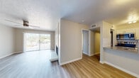 This single-floor unit offers a clean, modern aesthetic throughout, characterized by light-toned luxury vinyl plank flooring that runs continuously across the living, dining, and bedroom areas, creating a cohesive and visually expansive feel. Neutral beige walls and crisp white trim complement the textured popcorn ceilings, while a ceiling fan in the main living area promotes airflow and comfort.

The living space is bright and open, drawing natural light from a large sliding glass door fitted with vertical blinds that opens to a railed balcony with outdoor views of surrounding greenery. A half-wall partition separates the living area from the rest of the unit, adding subtle architectural definition without closing off the space.

The galley-style kitchen is well-equipped with stainless steel appliances, including an electric range, over-the-range microwave, and refrigerator. White shaker-style cabinetry, gray countertops, and a classic white subway tile backsplash give the kitchen a contemporary, polished look. A stainless steel sink completes the space.

The bedroom features a spacious closet with sliding panel doors, offering built-in shelving and a hanging rod for organized storage. The bathroom includes a full tub-shower combination, a white vanity with gray countertop, and wood-style flooring. The entry area provides a welcoming foyer with a windowed front door and ample wall space.
