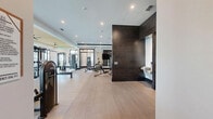 Located at Ascend Village in Winter Garden, Florida, this generously sized fitness center delivers a premium workout experience across a single, open floor plan. The space is thoughtfully designed with a contemporary aesthetic, featuring light wood-style flooring, crisp white walls accented by bold dark tile columns, and a distinctive yellow tray ceiling detail that adds a vibrant architectural focal point.

The main exercise area is expansive and open, housing a comprehensive array of strength training equipment including multi-use cable machines, functional training rigs, adjustable benches, a full dumbbell rack, and resistance accessories such as stability balls and resistance bands. A dedicated cardio zone lines the perimeter, equipped with treadmills and elliptical machines positioned in front of large, black-framed windows that frame lush views of tropical landscaping and a nearby pool, flooding the space with natural light.

A glass-enclosed section provides a mirrored training studio ideal for stretching or group workouts. Ceiling fans help maintain comfort alongside a modern HVAC system. A well-appointed service alcove includes a dual water fountain station, a refreshment counter, and cubbies for personal storage. The facility also includes a restroom for added convenience. Recessed lighting throughout ensures bright, even illumination during any time of day.