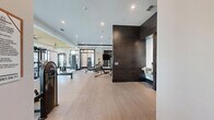 Located at Ascend Village in Winter Garden, Florida, this generously sized fitness center delivers a premium workout experience across a single, open floor plan. The space is thoughtfully designed with a contemporary aesthetic, featuring light wood-style flooring, crisp white walls accented by bold dark tile columns, and a distinctive yellow tray ceiling detail that adds a vibrant architectural focal point.

The main exercise area is expansive and open, housing a comprehensive array of strength training equipment including multi-use cable machines, functional training rigs, adjustable benches, a full dumbbell rack, and resistance accessories such as stability balls and resistance bands. A dedicated cardio zone lines the perimeter, equipped with treadmills and elliptical machines positioned in front of large, black-framed windows that frame lush views of tropical landscaping and a nearby pool, flooding the space with natural light.

A glass-enclosed section provides a mirrored training studio ideal for stretching or group workouts. Ceiling fans help maintain comfort alongside a modern HVAC system. A well-appointed service alcove includes a dual water fountain station, a refreshment counter, and cubbies for personal storage. The facility also includes a restroom for added convenience. Recessed lighting throughout ensures bright, even illumination during any time of day.