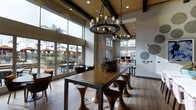 This single-story communal space is defined by its dramatic double-height ceilings accented with dark exposed beams and warm wood paneling, creating an open and airy environment. Two striking wrought-iron chandeliers fitted with vintage-style Edison bulbs serve as centerpiece fixtures, casting a warm, ambient glow throughout the room.

The main gathering area features a substantial communal high-top table in rich wood tones, surrounded by sculptural hourglass-shaped wooden stools, providing a casual yet stylish setting for socializing or working. The flooring throughout is light-toned hardwood, contributing to the warm aesthetic.

Floor-to-ceiling sliding glass doors and oversized windows along one wall flood the interior with natural light while offering direct visual access to an outdoor patio area, where seating and mature palm trees are visible. This seamless indoor-outdoor connection enhances the overall sense of openness.

Toward the rear of the space, a workstation or computer area is visible, suggesting a multi-functional design. Wall-mounted decorative elements and carefully placed greenery add finishing touches to the polished, contemporary ambiance of this well-appointed communal area.