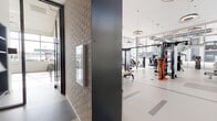 Located at 4109 E Tenth Ave in Denver's vibrant East Colfax neighborhood, this single-floor fitness center offers a well-appointed and thoughtfully designed workout environment. The space is defined by its dramatic floor-to-ceiling glass curtain walls, which flood the interior with abundant natural light and provide open views of the surrounding urban landscape. High ceilings with suspended round pendant lighting add to the airy, expansive feel of the facility.

The main workout floor is equipped with a comprehensive selection of Cybex strength machines, including selectorized and plate-loaded units with premium tan leather upholstery, as well as a multi-functional cable station. Free weight offerings include a full dumbbell rack and various benches. Cardio equipment — including treadmills, ellipticals, and stationary spin bikes — is strategically positioned along the windowed perimeter, allowing users to enjoy natural light and outside views during their workouts. Resistance training accessories such as TRX suspension trainers and resistance bands are neatly organized on a dedicated storage rack.

The facility features textured light-toned brick accent walls, durable resilient flooring with inlaid pattern detailing, and full-length mirrors that visually expand the space. A dedicated entry vestibule with glass doors and clean transitional finishes provides a polished arrival experience. The overall design balances industrial and modern aesthetics, creating a professional and motivating fitness environment.