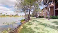 This single-story property is defined by its exceptional outdoor setting, positioned directly along the edge of a serene pond that serves as a defining feature of the surrounding landscape. The grounds are well-maintained, with a broad expanse of lush green lawn extending to the water's edge, bordered by a natural shoreline dotted with aquatic vegetation, including lily pads and native grasses. Mature trees, including tall cypress and palm varieties characteristic of Florida's natural environment, line the waterfront and provide generous shade across the grounds.

The building itself features a warm earth-toned exterior consistent with the Mediterranean-influenced architectural style commonly found in Florida residential communities. The property is part of a larger complex, with additional structures visible along the tree-lined waterfront, all sharing the same cohesive aesthetic. The reflective pond stretches broadly across the landscape, offering open water views framed by palms and distant greenery on the opposite shore.

The outdoor environment conveys a peaceful, resort-like ambiance, with natural elements playing a central role in the character of the property. The combination of open water, mature landscaping, and manicured lawn creates a tranquil setting well-suited to outdoor relaxation and enjoyment of the surrounding natural scenery.