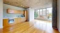 Located in the heart of Portland, Oregon, Unit 516 at 1720 SW 4th Ave presents a compelling urban loft aesthetic defined by its signature exposed concrete ceilings, which run throughout the main living areas and hallway, lending the space an industrial yet refined character. Warm hardwood flooring flows through the open-plan living and kitchen area, contrasting beautifully with the raw concrete overhead.

The kitchen is outfitted with warm-toned wood cabinetry, an under-counter oven, and a sink, all arranged in a clean galley configuration. Track lighting mounted to the concrete ceiling provides practical illumination. The living area benefits from expansive floor-to-ceiling windows that frame views of the surrounding Portland cityscape, including neighboring rooftops and greenery, while filling the interior with generous natural light.

A sliding frosted-glass panel separates the bedroom from the main living space, offering privacy without sacrificing light. The bedroom features carpet flooring, exposed concrete walls, and access to the building's exterior window wall. The bathroom is cleanly finished with a full tub-shower combination, tile flooring, and brushed nickel fixtures. A narrow hallway connects the rooms, with an exposed concrete accent wall adding to the unit's consistent industrial design language.