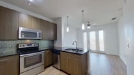 This thoughtfully designed one-bedroom, one-bathroom unit offers a functional and modern layout across a single floor. The open-concept kitchen and living area forms the heart of the unit, where the kitchen stands out with warm wood-toned cabinetry, a dark stone countertop, a mosaic tile backsplash, and a full suite of stainless steel appliances including a range, microwave, and dishwasher. A peninsula with an integrated sink and pendant lighting above creates a natural divide between the kitchen and the living space, which benefits from large French doors fitted with blinds that invite generous natural light and a ceiling fan overhead. Light wood-style flooring ties the common areas together seamlessly. The bedroom is a comfortable, carpeted retreat featuring large windows with blinds that allow for natural light while maintaining privacy, set against clean white walls. The bathroom is finished with large-format neutral tiles in the tub/shower surround, a soaking tub, a curved chrome shower rod, and matching wood-toned cabinetry with a dark countertop. Throughout the unit, the clean white walls and consistent modern finishes create a bright, cohesive, and contemporary ambiance.