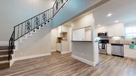 This two-story home showcases a thoughtfully designed layout with consistent, modern finishes throughout. The main level opens into a spacious living area with soaring ceilings and light-toned hardwood-style flooring that extends seamlessly across the space. A decorative staircase with ornate black metal balusters and white-painted steps serves as a striking architectural focal point, connecting the two floors with both style and function.

The open-concept kitchen is well-equipped with stainless steel appliances, including a range, microwave, dishwasher, and refrigerator. White shaker-style cabinetry pairs with dark countertops and a classic tile backsplash, creating a clean, contemporary aesthetic. Recessed lighting illuminates the kitchen, which benefits from a window that brings in natural light. A half-wall peninsula provides a natural separation between the kitchen and living areas.

Adjacent to the main living space, a built-in nook with white cabinetry offers additional storage and utility. The upper level hosts a well-sized bedroom featuring darker hardwood-style flooring, a ceiling fan, a window with blinds, and a walk-in closet with built-in shelving. The bathroom is finished with white cabinetry, a single-sink vanity with a dark countertop, a tub-shower combination, and matching hardwood-style flooring. The overall ambiance is bright, clean, and move-in ready.
