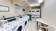This shared laundry facility is a practical, single-level space designed to accommodate multiple users efficiently. The room is equipped with several front-loading commercial washers and stacked dryer units, providing ample capacity for residents. A utility sink is available along one wall, offering added convenience for handwashing or pre-treating garments. Bright fluorescent overhead lighting ensures the space is well-lit throughout.

The interior features light tile flooring and cream-colored walls accented by a teal wainscoting detail that runs along the lower portion of the walls, lending a consistent visual identity to the room. A wall-mounted air conditioning unit helps maintain comfortable temperatures during use.

A folding table and seating area provide a functional spot for residents to manage laundry. A waste bin near the entrance keeps the space tidy. Safety is addressed through a clearly mounted fire extinguisher and posted emergency contact information near the teal entry door, which features a window allowing natural light to filter in. A posted laundry room rules sign outlines guidelines to ensure the facility remains clean and accessible for all users.