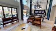 This sophisticated clubhouse lounge is designed to impress, combining dramatic architectural elements with a curated selection of social amenities. The space is anchored by soaring double-height ceilings that amplify the sense of openness, with large arched floor-to-ceiling windows and wide sliding glass doors that flood the interior with natural light and provide direct access to an outdoor pool area. The herringbone-patterned light wood flooring runs throughout, lending a refined yet relaxed aesthetic.

The main lounge area features rich cognac leather seating arranged around a gold-toned circular coffee table atop a blue and gold area rug, creating an inviting gathering space. A statement botanical chandelier and cascading pendant globe lights serve as artistic focal points suspended high overhead.

A full wet bar with a sleek white quartz countertop and dark cabinetry offers a dedicated space for entertaining, complemented by multiple wall-mounted screens above a tiled backsplash. Adjacent to the lounge, a shuffleboard table and an oversized wall-mounted Scrabble board set into a dark textured accent wall provide interactive entertainment options. Floor-to-ceiling teal drapery panels add a dramatic yet elegant touch, subtly dividing zones while maintaining visual flow throughout the space.