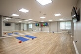 Located at Avalon Square in Glen Cove, New York, this single-floor fitness and yoga studio is thoughtfully designed to support a wide range of exercise activities. The open layout features smooth hardwood-style flooring throughout, providing an ideal surface for yoga, stretching, and group fitness sessions. A large floor-to-ceiling mirror spans one full wall, enhancing the sense of space while allowing users to monitor their form during workouts. The studio is well-lit by recessed panel lighting in the ceiling, supplemented by natural light streaming through windows fitted with adjustable blinds. The neutral gray walls and clean white trim create a calm, focused atmosphere. Equipment includes exercise mats, hand weights, kettlebells, and a collection of stability balls mounted on wall racks, offering variety for both cardio and strength training. A wall-mounted flat-screen television is positioned for easy viewing during workouts. The space also includes a coat hook wall rack, in-ceiling speakers, and a separate entry door providing convenient access. A built-in HVAC unit ensures year-round comfort. Interior windows connect the studio to an adjacent fitness area, adding visibility and a sense of connectivity throughout the facility.