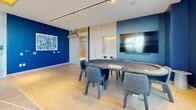 Located on Rathburn Road West in Mississauga, Ontario, this single-floor entertainment and games lounge is a thoughtfully designed amenity space that combines vibrant aesthetics with a variety of recreational offerings. The space is defined by its bold navy and cobalt blue accent walls, warm light wood flooring, and a striking yellow-backlit tray ceiling that adds a dramatic flair to the games area.

A full-size pool table anchors one section of the room, suspended beneath a sleek linear pendant light and set against a large-format stone-clad wall with a wall-mounted flat-screen television. Floor-to-ceiling windows flood this area with abundant natural light while framing views of the surrounding Mississauga cityscape. A dedicated poker table with upholstered seating occupies an adjacent zone, accompanied by a wall-mounted display and built-in light wood cabinetry with integrated storage.

The lounge area offers a more relaxed atmosphere, featuring a circular wooden coffee table, woven accent chairs, and a comfortable sofa arranged on a colorful geometric area rug. A marble-textured feature wall serves as a backdrop for another large flat-screen television, completing the media setup. Track lighting throughout ensures versatile illumination across all zones. The cohesive design palette and multi-functional layout make this an inviting destination for social gatherings and leisure activities.