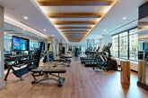 Located at 285 Mayson Ave NE in Atlanta, GA, this expansive fitness center offers a thoughtfully designed, single-floor layout spanning a generous footprint. The space strikes a refined balance between functionality and modern aesthetics, making it a standout facility for health and wellness.

The architectural design is defined by a striking coffered ceiling adorned with warm wood beams and recessed lighting that casts an inviting, ambient glow throughout. Wide-plank hardwood flooring runs the length of the space, lending a clean and upscale feel underfoot. Large floor-to-ceiling windows line the perimeter, flooding the interior with natural light and offering views of the surrounding outdoor greenery.

The fitness floor is comprehensively equipped, featuring a full row of treadmills, elliptical machines, and stationary bikes for cardio training, alongside an array of cable machines, multi-use strength stations, free weights, and adjustable benches for resistance training. Black steel room dividers provide subtle zoning throughout the open floor plan without interrupting the sense of spaciousness.

Full-length mirrors are strategically positioned to enhance the sense of openness and support proper form during workouts. A dedicated hydration and amenity station, finished with wood cabinetry and decorative tilework, adds a polished, hospitality-inspired touch. Digital display screens are integrated throughout, providing on-demand fitness content and programming information.