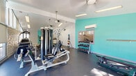Located at 9550 Fremont Ave in Montclair, California, this fitness center offers a comprehensive workout facility on a single floor. The space is thoughtfully designed with a vaulted ceiling, pendant ceiling fans, and recessed fluorescent lighting that creates a bright and energizing atmosphere.

The exercise room is finished with durable speckled rubber flooring and features a vibrant teal accent wall that adds a modern, energetic feel to the space. Climate comfort is ensured by a wall-mounted split AC unit, making the facility usable year-round.

Equipment is well-distributed throughout the room and includes a multi-station cable weight machine, elliptical trainers, a StairMaster stepmill, a treadmill, and an elliptical cross-trainer. A dedicated free weights area houses a fully stocked dumbbell rack alongside a selection of medicine balls, and a weight bench provides additional strength training options. A wall-mounted mirror enhances the sense of space and supports proper form during workouts.

Large sliding glass doors and expansive windows along one wall flood the space with natural light and offer direct views of an outdoor pool and patio area, creating a refreshing connection to the outdoors. White brick-textured walls add architectural character, while roller shades on the windows allow for adjustable light control. The overall ambiance is clean, functional, and motivating.