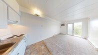 This single-floor unit features a bright, open layout that seamlessly connects the living area and kitchen. The main living space is finished with neutral carpet and crisp white walls, creating a clean and airy atmosphere. Paneled ceilings add a subtle architectural detail overhead, while baseboard heaters run along the walls, providing consistent warmth throughout. A wall-mounted air conditioning unit ensures year-round comfort. A sliding glass door at the far end of the living area floods the space with natural light and provides direct access to a balcony, offering pleasant outdoor views of surrounding greenery.

The kitchen area is tucked neatly into one corner and features white cabinetry with ample upper and lower storage, a granite-style countertop, a stainless steel sink, and a white electric coil range with an overhead exhaust hood. The kitchen is finished with durable tile flooring, contrasting with the carpeted living area.

The bathroom is clean and functional, featuring a vanity with white cabinetry, a patterned tile floor, a toilet, and baseboard heating. White walls and a louvered cabinet door contribute to the room's tidy, organized appearance.