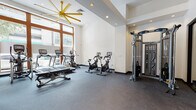 Located at 2081 Center St in Berkeley, CA, this fitness center offers a comprehensive, single-floor workout facility designed to support a wide range of exercise routines. The space is defined by clean white walls, durable rubber flooring, and a bright, open layout that promotes ease of movement throughout.

Natural light pours in through large wood-framed windows, creating an inviting and energizing atmosphere. A striking yellow starburst ceiling fan serves as a bold architectural accent, complementing the recessed lighting overhead and adding a distinctive design element to the space.

The cardio zone is well-stocked with multiple treadmills, stationary bikes with integrated display screens, and an elliptical machine, all arranged to take advantage of the natural light. Adjacent to this area, a multi-station cable machine provides options for a full range of strength training exercises.

A dedicated free weights section features a tiered dumbbell rack with a wall mirror, allowing users to monitor their form during workouts. The functional training area includes a heavy bag, TRX suspension trainers, battle ropes, resistance tools, and plyo boxes, offering versatile cross-training options.

A corridor near the entrance provides a dual water fountain station for hydration convenience, alongside restroom access. The overall layout is thoughtfully organized, balancing cardio, strength, and functional fitness within a cohesive and well-maintained environment.