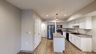 This single-floor residence is designed with a clean, contemporary aesthetic and consistent use of wood-look plank flooring that runs throughout the space. The kitchen serves as a focal point, showcasing white shaker-style cabinetry, granite-look countertops, and a full suite of stainless steel appliances, including a refrigerator, electric range, microwave, and dishwasher. A center island provides additional prep and casual dining space, while recessed lighting and a decorative pendant fixture illuminate the area effectively. The kitchen also features an undermount sink and ample upper and lower cabinet storage.

The layout, as shown in the 3D overview, includes multiple rooms branching off a central corridor. The bedrooms are finished with the same wood-look flooring, neutral gray walls, and feature windows dressed with horizontal blinds that allow natural light in. One bedroom includes a ceiling fan and a spacious closet with a paneled door. The bathroom is equipped with a walk-in shower unit featuring built-in shelving niches and a curtain rod. Throughout the unit, neutral gray walls, white trim, and consistent finishes create a cohesive, modern atmosphere.