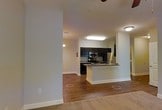 This single-floor residence offers a thoughtfully designed open-concept layout that connects the living and kitchen areas in a seamless flow. The living room features soft carpeting, neutral gray-toned walls, and a ceiling fan, creating a comfortable and relaxed atmosphere. A partial dividing wall adds subtle definition between spaces without sacrificing the open feel of the interior.

The kitchen area is positioned adjacent to the living space and showcases a granite-style countertop that adds a touch of elegance to the functional layout. The transition from carpet to rich, hardwood-style flooring marks the shift between the living and kitchen zones, adding warmth and visual contrast throughout the space.

A hallway extends toward the back of the residence, leading to additional rooms. The corridor is well-lit with overhead lighting and features the same warm hardwood-style flooring, maintaining a cohesive aesthetic. White baseboards and trim offer clean, crisp accents against the softly painted walls throughout.

Overall, the space presents a clean, move-in ready environment with a neutral palette that provides a versatile backdrop for a variety of interior styles. The practical layout and consistent finishes give the residence a welcoming and well-maintained character.