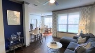This single-floor residence showcases an open-concept design that integrates the living, dining, and kitchen areas into one cohesive, flowing space. The living area features dark hardwood-style flooring that extends throughout, grounding the space with a warm, contemporary aesthetic. A ceiling fan with a light fixture overhead provides both comfort and ambient illumination.

A breakfast bar with white upholstered high-back stools serves as a natural divider between the living space and the kitchen, offering a casual dining option while maintaining the open feel. Track lighting runs along the ceiling, providing directed illumination across the layout.

The dedicated dining area is positioned near large windows dressed with patterned curtains and horizontal blinds, allowing generous natural light to fill the space while offering views of the surrounding greenery outdoors. The dining setup accommodates multiple guests comfortably.

Architectural details such as a decorative column near the bar area and gray crown molding add a polished finish to the interiors. The overall ambiance is bright, modern, and welcoming, with a clean, neutral palette that allows for versatile styling throughout.