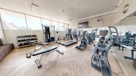 Located at 6303 Indian School Rd NE in Albuquerque, New Mexico, this fitness center offers a well-appointed and efficiently organized workout space on a single floor. The room is bathed in natural light streaming through large, floor-to-ceiling windows that line the walls, providing open views of the surrounding Albuquerque landscape and creating a bright, energizing atmosphere. The flooring features a light wood herringbone pattern that adds a clean, modern aesthetic throughout.

The space is comprehensively equipped to support a full range of workout routines. Cardio equipment includes multiple treadmills, elliptical trainers, and stair-climbing machines, all arranged to take advantage of the natural light and outdoor views. A Hoist functional training cable machine serves as a centerpiece for strength and functional fitness, complemented by a multi-station weight machine and a full rack of dumbbells ranging across various weights. Adjustable workout benches are positioned throughout for versatile strength training exercises.

A wall-mounted flat-screen television provides entertainment during workouts, while a wall clock keeps exercisers on schedule. Vertical blinds on select windows offer adjustable light control. The overall ambiance is clean, open, and functional, making this fitness center a practical and inviting space for residents or guests seeking a complete workout facility.