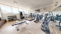 Located at 6303 Indian School Rd NE in Albuquerque, New Mexico, this fitness center offers a well-appointed and efficiently organized workout space on a single floor. The room is bathed in natural light streaming through large, floor-to-ceiling windows that line the walls, providing open views of the surrounding Albuquerque landscape and creating a bright, energizing atmosphere. The flooring features a light wood herringbone pattern that adds a clean, modern aesthetic throughout.

The space is comprehensively equipped to support a full range of workout routines. Cardio equipment includes multiple treadmills, elliptical trainers, and stair-climbing machines, all arranged to take advantage of the natural light and outdoor views. A Hoist functional training cable machine serves as a centerpiece for strength and functional fitness, complemented by a multi-station weight machine and a full rack of dumbbells ranging across various weights. Adjustable workout benches are positioned throughout for versatile strength training exercises.

A wall-mounted flat-screen television provides entertainment during workouts, while a wall clock keeps exercisers on schedule. Vertical blinds on select windows offer adjustable light control. The overall ambiance is clean, open, and functional, making this fitness center a practical and inviting space for residents or guests seeking a complete workout facility.