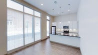This single-floor, one-bedroom, one-bathroom unit showcases a contemporary open-concept design that maximizes its well-proportioned layout. The living and kitchen area is immediately distinguished by soaring ceilings and expansive floor-to-ceiling windows fitted with horizontal blinds, flooding the space with natural light while offering views of the surrounding streetscape. Warm wood-look flooring flows throughout the main living area, creating a cohesive and inviting atmosphere.

The kitchen is a standout feature, centered around a large peninsula with a dark granite countertop that provides ample prep and seating space. White shaker-style cabinetry lines the walls, complemented by under-cabinet lighting and a full suite of stainless steel appliances, including a refrigerator, range, and microwave. Three sleek pendant lights hang above the peninsula, adding a polished finishing touch.

The bedroom offers a quieter retreat, finished with plush neutral carpeting and tall ceilings that lend a sense of openness. It connects directly to the full bathroom, which features a tub-shower combination, wood-look flooring, a dark granite vanity top, and clean white walls that contribute to the unit's consistent modern aesthetic throughout.