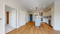 This well-appointed single-floor space showcases a thoughtfully designed open-concept layout that seamlessly connects the living and kitchen areas. The kitchen is a standout feature, outfitted with crisp white shaker cabinetry, a subway tile backsplash, and a full suite of stainless steel appliances including a French door refrigerator, range with wall-mounted hood vent, and dishwasher. A central island with a veined quartz countertop serves as both a functional workspace and a visual anchor, accented by dual pendant lights overhead. Warm-toned hardwood flooring flows throughout the main living areas, creating a cohesive and inviting atmosphere. Recessed lighting throughout ensures the space remains bright and well-lit. The bedroom offers a change in texture with plush carpet flooring and features a large window that frames an outdoor water view, filling the room with natural light. The bathroom is finished in a clean, minimalist style with large-format tile flooring, a full soaking tub with overhead shower, and a sleek white floating vanity. Multiple interior doors and a distinctive teal entry door add subtle character to the otherwise neutral palette, giving the space a modern yet welcoming feel.