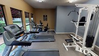 This fitness center offers a practical and organized workout environment spread across a single floor. The space features a two-toned interior, with warm tan walls accented by cool gray panels, complemented by a textured ceiling with flush fluorescent lighting throughout. Carpet flooring covers the entire space, providing a comfortable surface underfoot.

The cardio area is anchored by multiple Matrix-brand treadmills lined along the windowed wall, which allows natural light to filter in and offers views of the surrounding greenery outdoors. Alongside the treadmills sit an upright stationary bike and a recumbent bike, rounding out the cardiovascular equipment selection.

On the opposite side of the room, a white multi-station cable weight machine serves as the centerpiece for strength training, accompanied by adjustable incline benches for a variety of exercises. A sanitizing wipe dispenser and waste bin are conveniently positioned near the entry door, promoting cleanliness and hygiene. A posted fitness center rules sign is prominently displayed on the wall, ensuring all users are informed of facility guidelines.

Access to the fitness center is provided through a glass-paneled entry door, which connects to an exterior walkway surrounded by lush landscaping. The overall layout is efficiently arranged to maximize usability while maintaining an open, uncluttered feel throughout the space.