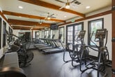 Located at 470 Saratoga Ave in San Jose, CA, this fitness center offers a thoughtfully designed workout environment across two exercise rooms on a single floor. The space is characterized by soaring ceilings with rich, exposed wooden beams that lend a warm, rustic-modern aesthetic to the otherwise crisp white walls and dark-trimmed black windows. Rubber flooring throughout ensures durability and safety during workouts.

The main exercise room is generously outfitted with a wide array of cardio equipment, including treadmills, elliptical machines, and stationary bikes, all arranged to maximize floor space and usability. A wall-mounted television provides entertainment during workouts, while ceiling fans maintain comfortable airflow. Full-length mirrors along one wall enhance the sense of openness and allow users to monitor their form.

The second exercise area features strength training equipment, including a multi-functional cable machine, a dumbbell rack with a full set of free weights, and an adjustable weight bench. Large black-framed windows flood both rooms with natural light, offering views of the surrounding greenery. The architectural detail of a dramatic arched window adds a distinctive visual element to the space. The overall ambiance is clean, functional, and inviting, making it a well-rounded facility for a comprehensive fitness routine.