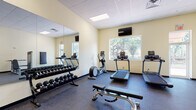 This fitness center at 5500 Main St in New Port Richey, Florida, offers a well-equipped, single-floor facility designed to support a full range of workout routines. The space is thoughtfully organized into distinct exercise zones, all unified by warm cream-colored walls, a drop-panel ceiling with recessed lighting, and durable rubber flooring throughout.

The cardio area is outfitted with multiple treadmills, an elliptical trainer, and a step machine, arranged to face a large picture window that provides views of the surrounding greenery — creating a bright and inviting atmosphere. A wall-mounted flat-screen television adds to the user experience during cardio sessions.

The strength training section features a full rack of dumbbells in varying weights, complemented by a large wall mirror that enhances the sense of space and allows for proper form checks. Several cable and selectorized weight machines, including a lat pulldown, leg curl, and chest press, provide comprehensive muscle group targeting.

A dedicated area accommodates stability balls and functional training accessories, offering flexibility for stretching and core work. Natural light filters in through multiple windows and glass-panel doors, connecting the interior to the outdoors. The facility presents a clean, functional, and professional environment suitable for users of all fitness levels.