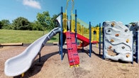 Located at 7999 Wildflower Ln in Westerville, Ohio, this outdoor recreational space offers a well-equipped playground designed to engage children of various ages and abilities. The centerpiece of the space is a colorful, multi-level play structure featuring bold navy blue, yellow, and red components that stand out against the lush green surroundings. Children can explore a variety of activity zones, including a twisting tube slide, a rock climbing wall with textured handholds, a rope ladder climbing panel, and a staircase with bright yellow railings leading to an elevated platform. The structure is thoughtfully designed to encourage active play and physical development through multiple access points and activity options. The ground beneath the equipment is covered with a rubber mulch safety surface, providing cushioning for active play. The surrounding landscape features open grassy areas with mature trees offering natural shade, making it a welcoming environment on sunny days. The expansive open lawn surrounding the playground provides additional space for unstructured outdoor activities. The overall setting is bright, inviting, and well-maintained, making this playground a functional and engaging destination for the local community.