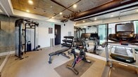 Located at 3800 Mentone Ave in Culver City, California, this well-appointed fitness center showcases an industrial-modern aesthetic defined by exposed concrete ceilings with visible ductwork, recessed track lighting, and a polished epoxy-style floor. The single-level space is thoughtfully organized into distinct training zones that together accommodate a comprehensive range of workout styles.

The cardio zone features multiple treadmills and elliptical machines, each equipped with individual screens, and is positioned along a mirrored wall that amplifies the sense of space and allows users to monitor their form. An adjacent strength training area includes a full cable and multi-function rack system, a tiered dumbbell rack with a wide weight selection, adjustable benches, and a spin bike, providing versatile options for resistance and interval training.

Large sliding glass doors open directly onto an outdoor turf terrace, where stability balls and additional equipment are accessible, bringing natural light deep into the interior and creating a seamless indoor-outdoor training experience. Views of surrounding greenery are visible through both the terrace and interior windows, enhancing the overall ambiance. The space is branded with MV signage and maintains a clean, professional atmosphere suited to serious training.