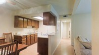 This single-floor space presents a practical and straightforward layout, centered around a kitchen that is both functional and well-appointed. The kitchen features rich, dark wood cabinetry lining the walls, providing ample storage alongside a countertop with an integrated sink. A peninsula-style cooktop serves as a natural divider, separating the kitchen from the adjacent hallway and adding an element of openness to the layout. Overhead, a suspended dark wood cabinet adds additional storage while contributing to the kitchen's defined character. Light-toned laminate flooring runs consistently through the kitchen and into the hallway, creating visual continuity throughout the space. A skylight-style ceiling fixture brightens the kitchen area with additional illumination. The hallway extending from the kitchen is clean and well-lit, leading to further rooms within the unit, suggesting a straightforward and efficiently organized floor plan. Neutral wall tones and standard ceiling height maintain a calm, understated ambiance throughout. The HVAC vent integrated into the ceiling indicates climate-controlled comfort throughout the space.