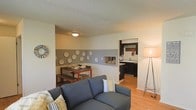 This single-floor residence offers an open and functional layout that seamlessly connects the living, dining, and kitchen areas. The living room is anchored by a grey sofa accented with patterned throw pillows, complemented by warm wood-style flooring that extends throughout the space. A tripod floor lamp adds a modern touch, while a decorative sunburst mirror adorns one wall, contributing to the room's casual yet curated feel.

The dining area is defined by a rich wood table paired with a matching bench and chairs, set against a two-tone accent wall in grey and white. Decorative circular plates are artfully arranged on the wall, adding visual interest and a personalized touch. A pass-through opening connects the dining space to the kitchen, enhancing the open-concept flow.

The kitchen features dark cabinetry contrasted against light countertops, with a sink positioned beneath a mirror that reflects natural light into the space. Wood-style flooring continues from the main living area into the kitchen, maintaining a consistent design language. A textured ceiling is present throughout, and flush-mount lighting provides adequate illumination across the unit.